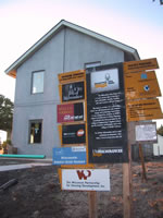 This photograph shows the construction site of the Milwaukee Idea Home with the building structure completed on it in the background and a sign explaining what it is, the project partners, and program sponsors in the foreground.