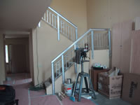 This photograph, taken from the first floor, shows a near-completed stairway during the construction of the Milwaukee Idea Home. The stairway zigzags from the first floor to the second floor. The stairway extends upwards from the first floor to the right where it meets a landing and then switches back to the left before it reaches the second floor. The viewer is able to see how sturdy and well constructed the railings on the sides of the stairway will be and what the stairway looks like. Tools, construction equipment, boxes, and a protective floor covering surround the stairway.