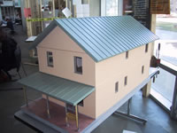 The photograph shows the exterior of a model house display. The model is fairly large and spans the entire length of a rectangular table top. The roof, first and second floors and a porched-in front section of the house are featured in the photo. Several opaque windows are also seen on the first and second floors of the house.