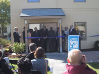The photograph displays a celebratory ribbon-cutting ceremony for the completion of the Milwaukee Idea Home. The owner and those in charge of the project are featured in this photograph and are seconds away from cutting a ribbon that symbolizes that the house has been completed. The ribbon is in front of the backside of the model house and attached to the small porch area by the back door. An audience has gathered to observe the event. A large Independence First sign is attached to a portion of the ribbon advertising the ownership of the organization that directed the project.