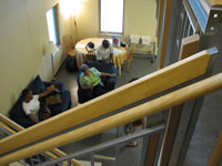 The photograph shows a tour being conducted inside of the model house. The view is from the top of the stairway and the camera is aimed downwards displaying the living room and several people within it. The people appear to be listening to a person, most likely a tour guide who is not shown in the photograph. The view that the camera angle creates from the second floor, gives the viewer a unique portrait of the lower level room and the basic lay-out of the room. The room includes a couch, a chair, a kitchen or dining room table, a couple windows and lamps and a rolling cart.