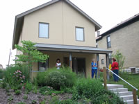 This photograph shows a tour of the exterior of the completed Milwaukee Idea Home. Four people are in the front of the house looking around. The front lawn, first and second floors, and pathway with a staircase are visible in the photograph. One of the persons in the photograph appears to be the tour guide and she is speaking with an older lady.