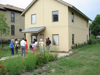 The photograph shows a tour being given outside of the model home. The view is from the backside of the house and the tour guide and members are standing around by the back door. The entire back and side of the house can be seen in the photograph as well as the back yard.