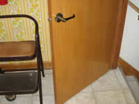 This photograph features a lever door handle that has been installed on a basement door. The lever door is large and designed to be universally accessible. The handle can be easily manipulated and does not require a great deal of fine motor control. It can be activated or turned with various body parts, assistive devices, or objects, in addition to a hand or fingers.