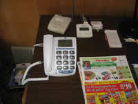 This photograph features a low-vision telephone that has been placed on top of a table. The phone is easily accessible for people with low vision. The phone features large gray buttons with big white numbers on them fostering a high contrast from the background, making it easier to see. There also is a display with large letters and numbers on the phone, so that the user is able to more easily identify who has called, whether there are any messages, and see the date and time. The phone is attached by a large white cord and plugged into a phone jack on the wall behind it.