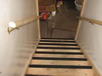 This photograph features railings on both sides of a basement stairwell and grip-tape on the edge of each step. The double railings are large and wooden, enabling increased balance and stability of the user. The black, abrasive strips attached on the ends of each step are slip-resistant. The strips are composed of a gritty, textured material that increases tread and traction on the wooden steps. This feature also increases safety and decreases the chances of falls occurring on the stairs. The strips comply with accessibility standards and provide a good safety precaution for homeowners of all ages.