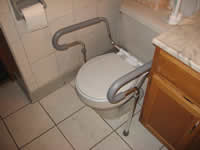 This photograph features large removable grab bars that have been attached on either side of a toilet. The grab bars are unique because they can be flipped up and temporarily positioned out of the way. The grab bars also are useful to enable proper body mechanics and to prevent falls or slips. The grab bars can easily be assembled and removed. Because the grab bars have a larger diameter, they are more easily grasped.