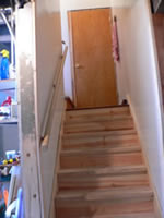 TThis photograph represents a stairway that leads from the main floor of a home to the basement. The stairs are made of unfinished wood and there is only one railing. An individual using the staircase is at greater risk of injury or falls due to the slippery wooden stairs, low contrast between the steps, and a single railing.