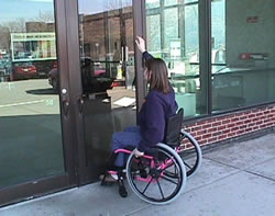 The woman in a wheelchair is trying to open a door to Enderis Hall on the UW-Milwaukee campus. Unfortunately, this entrance is not accessible.  Because there are no push buttons to assist the woman in opening the door, she is attempting to flag down a passerby for assistance.  This photograph depicts an example of advocacy.  There is a person in a wheelchair waving, yelling, and knocking at a door to draw attention to herself to let others know that she needs assistance to get into the building.  This image represents the sometimes limited accessibility resources on campus and the struggles a student may face when they encounter a similar situation.  The ACCESS-ed project focuses on campus accessibility to remove barriers and incorporate universal design on campus.